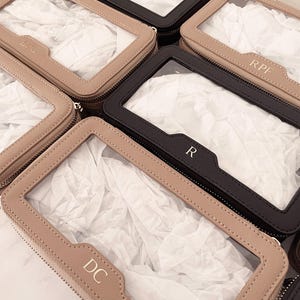 Puede incluir: Varias bolsas de cosm&eacute;ticos personalizadas con ventanas transparentes. Las bolsas son de color negro y beige, con cremalleras plateadas e iniciales en la parte delantera. Las bolsas est&aacute;n rellenas de papel de seda blanco.