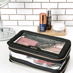 Puede incluir: Bolsa de cosm&eacute;ticos negra y transparente de doble capa con cremalleras doradas y las iniciales "MN". La bolsa contiene productos de maquillaje. Productos de belleza adicionales se muestran en una bandeja de madera.