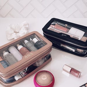 Puede incluir: Dos neceseres transparentes, uno beige y otro negro, llenos de productos de belleza. La bolsa beige est&aacute; etiquetada como "ANA". Tambi&eacute;n se ven un dispositivo Foreo rosa y un brillo labial. Tambi&eacute;n hay una goma para el pelo blanca y una paleta de maquillaje.