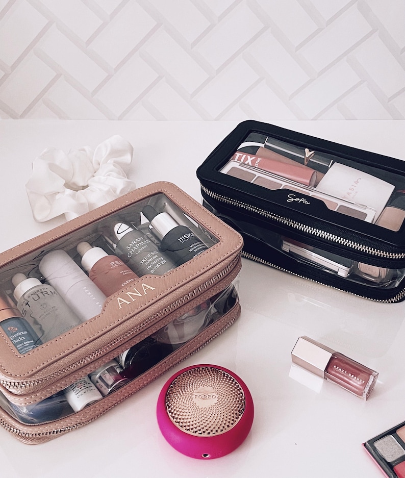 Puede incluir: Dos neceseres transparentes, uno beige y otro negro, llenos de productos de belleza. La bolsa beige est&aacute; etiquetada como "ANA". Tambi&eacute;n se ven un dispositivo Foreo rosa y un brillo labial. Tambi&eacute;n hay una goma para el pelo blanca y una paleta de maquillaje.