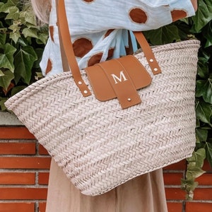 Op de afbeelding: Een witte geweven tote bag met bruine leren banden en een bruine leren flap met de letter "M" in wit gegraveerd.