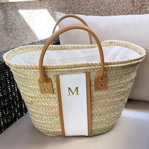 Puede incluir: Bolso tote de paja tejida con asas y detalles en cuero marrón. La bolsa presenta un panel de cuero blanco con la letra dorada "M". La bolsa tiene forma rectangular y está diseñada para transportar objetos.