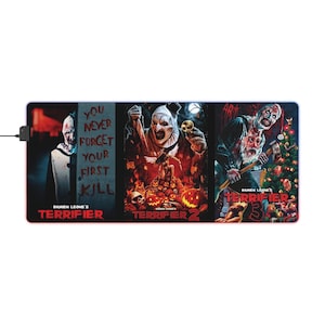 Puede incluir: Alfombrilla de ratón rectangular con tres paneles de películas de terror. Los paneles muestran un payaso y el texto "YOU NEVER FORGET YOUR FIRST KILL", "TERRIFIER 2" y "TERRIFIER 3". La alfombrilla tiene un borde negro y un conector USB.