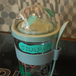 May include: A light blue plastic container with a clear lid and a built-in spoon. The container has a sticker that says "GRAPES" and a design of grapes on the lid.