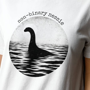 Puede incluir: Camiseta blanca con un gráfico en blanco y negro de un monstruo del Lago Ness con el texto "non-binary nessie" sobre el gráfico.