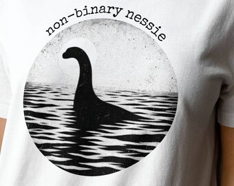 Può includere: Maglietta bianca con una grafica in bianco e nero di un mostro di Loch Ness con il testo "non-binary nessie" sopra la grafica.