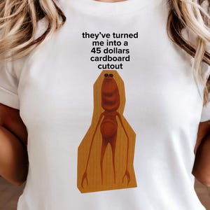 以下が含まれることがあります： 漫画のキャラクターのユーモラスなグラフィックと「they've turned me into a 45 dollars cardboard cutout」というテキストが入った白いTシャツ。このシャツはデニムショーツと組み合わせています。