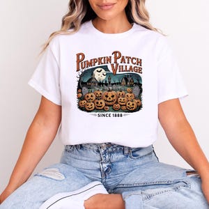 Könnte beinhalten: Weißes T-Shirt mit einem Halloween-Motiv. Das Design zeigt "Pumpkin Patch Village" über einer Szene mit Kürbissen, einem Vollmond und einem Spukhaus. Der Text "SINCE 1888" befindet sich unter der Szene.