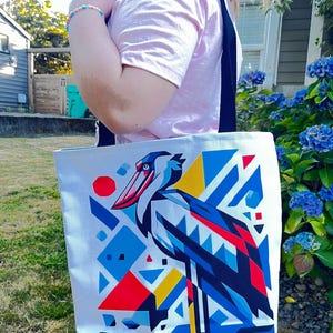 May include: A white tote bag with a colorful geometric design featuring a stylized shoebill stork. The design is in shades of blue, red, yellow, and black.