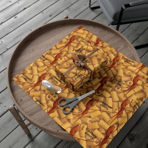 May include: A wrapped gift box and wrapping paper featuring a repeating pattern of rigatoni pasta and the words "rigatoni pasta" in a playful font. A pair of scissors and a spoon are on the paper.
