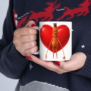 Puede incluir: Taza de cerámica blanca con un personaje de dibujos animados y un corazón rojo. El personaje es naranja con extremidades largas y gafas de sol. La taza la sostiene una persona que lleva un jersey azul oscuro.