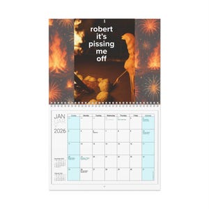 May include: A spiral-bound wall calendar for January 2026, featuring a humorous image with text that reads "robert it's pissing me off." The calendar has a blue and white grid for the days of the month, with a smaller calendar for December 2025.