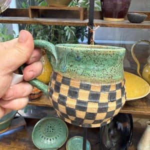 May include: A ceramic mug featuring a speckled light green glaze on the upper portion and a black and tan checkered pattern on the lower portion. The mug has a sturdy handle and a rounded body, showcasing a distinct contrast between the two textures.
