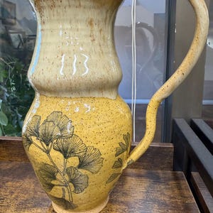 May include: A tall ceramic pitcher with a two-tone yellow glaze. The upper section is a pale cream-yellow, while the lower bulbous body is a deeper mustard yellow with dark speckles and a black line-art illustration of ginkgo biloba leaves.