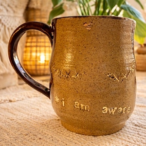 Puede incluir: Taza artesanal de cerámica de color beige con asa marrón oscuro. La taza presenta detalles cosidos y las palabras "i am aware" grabadas en la superficie. Una taza de café rústica y única para el uso diario.
