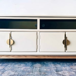 May include: A white media console with three doors and a shelf. The doors have gold circular handles. The console has a light wood base.