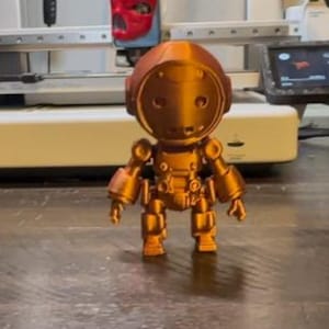 May include: A 3D printed, copper-colored robot figurine with a round head and a detailed body. The robot is standing on a wooden surface.
