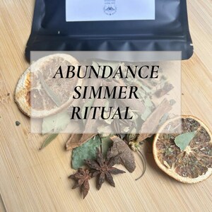 May include: A wooden surface displays dried botanicals, including star anise, orange slices, and bay leaves. A black pouch labelled "Abundance Simmer Pack" sits above the arrangement. The text "ABUNDANCE SIMMER RITUAL" is overlaid on the botanicals.