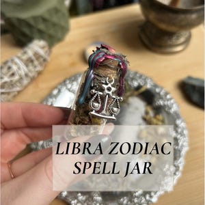 Peut inclure: Un petit bocal en verre rempli d'herbes et scellé avec de la cire. Le bocal porte un charme argenté avec un motif de balance. Le texte "LIBRA ZODIAC SPELL JAR" est affiché dans un encadré blanc. Le bocal est sur un plateau argenté.