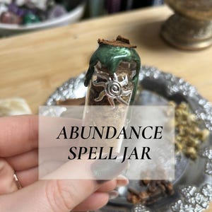 Pode incluir: Um frasco de vidro rotulado "ABUNDANCE SPELL JAR" preenchido com ervas e selado com cera verde e um amuleto de sol. O frasco está sobre uma bandeja de prata decorativa, sugerindo um tema espiritual ou metafísico.