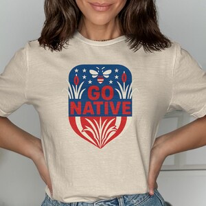 Puede incluir: Camiseta beige con un diseño patriótico. El gráfico presenta una forma de escudo con las palabras "GO NATIVE" en rojo, una parte superior azul con estrellas y una abeja, y detalles en rojo y blanco.