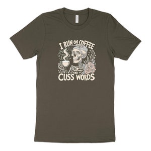 May include: A dark green t-shirt featuring a graphic of a skull drinking coffee and the text "I run on coffee and cuss words".