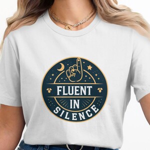 Könnte beinhalten: Weißes T-Shirt mit einem marineblauen Kreis-Motiv, das eine Hand mit einem nach oben zeigenden Finger, Sterne und einen Halbmond zeigt. Der Text "Fluent in Silence" ist in goldener Schrift geschrieben.