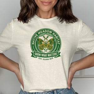 Cream t-shirt with green circular Native Meadow Habitat logo featuring butterflies and EST. 10,000 BCE.