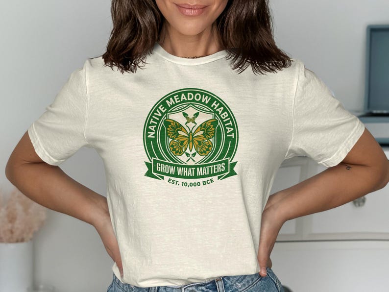 Cream t-shirt with green circular Native Meadow Habitat logo featuring butterflies and EST. 10,000 BCE.