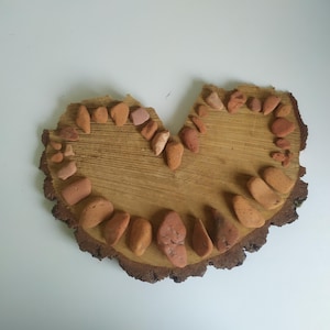Puede incluir: Una rodaja de madera en forma de corazón con un borde de pequeñas piedras de color marrón rojizo. Las piedras están dispuestas en forma de corazón, con un espacio en la parte superior.