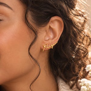 May include: A gold-toned ear climber earring featuring the name Raegan written in a cursive script font. The metal jewelry piece follows the curve of the earlobe, extending upwards with a polished finish and a delicate, thin wire construction.
