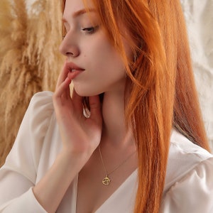 May include: A woman with long, red hair wearing a white blouse with a gold pendant necklace featuring a geometric design.