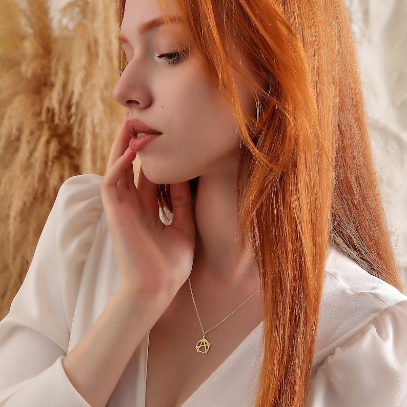 May include: A woman with long, red hair wearing a white blouse with a gold pendant necklace featuring a geometric design.