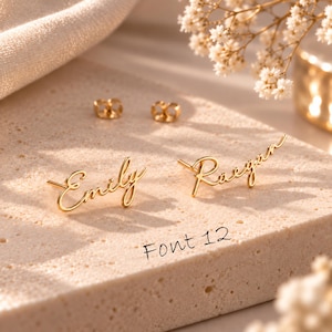 May include: A pair of gold-toned stud earrings featuring the names Emily and Raegan written in a cursive script font. The earrings include two matching gold-toned earring backs. The text Font 12 is printed on the surface near the earrings.