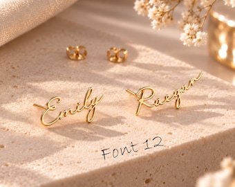 May include: A pair of gold-toned stud earrings featuring the names Emily and Raegan written in a cursive script font. The earrings include two matching gold-toned earring backs. The text Font 12 is printed on the surface near the earrings.