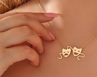 May include: A gold-toned necklace featuring a pendant shaped like the comedy and tragedy theater masks. The pendant is suspended from a delicate gold-toned chain, showcasing the iconic dual expression of a smiling face and a frowning face.