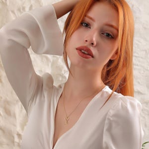 K&ouml;nnte beinhalten: Eine Frau mit langen, roten Haaren tr&auml;gt eine wei&szlig;e Bluse mit einer goldenen Halskette mit einem geometrischen Anh&auml;nger.