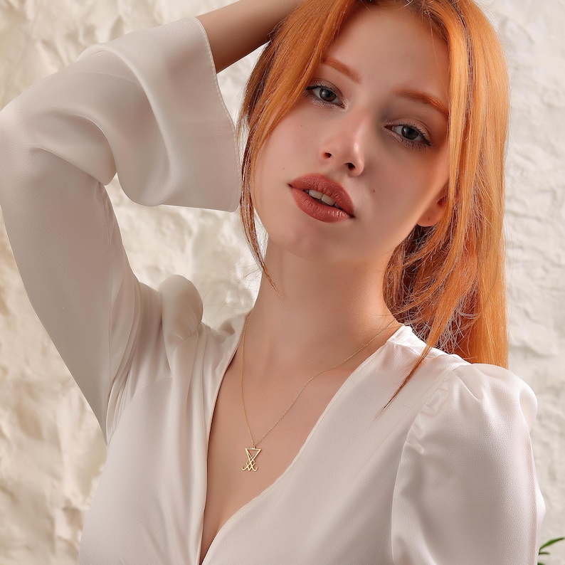 K&ouml;nnte beinhalten: Eine Frau mit langen, roten Haaren tr&auml;gt eine wei&szlig;e Bluse mit einer goldenen Halskette mit einem geometrischen Anh&auml;nger.