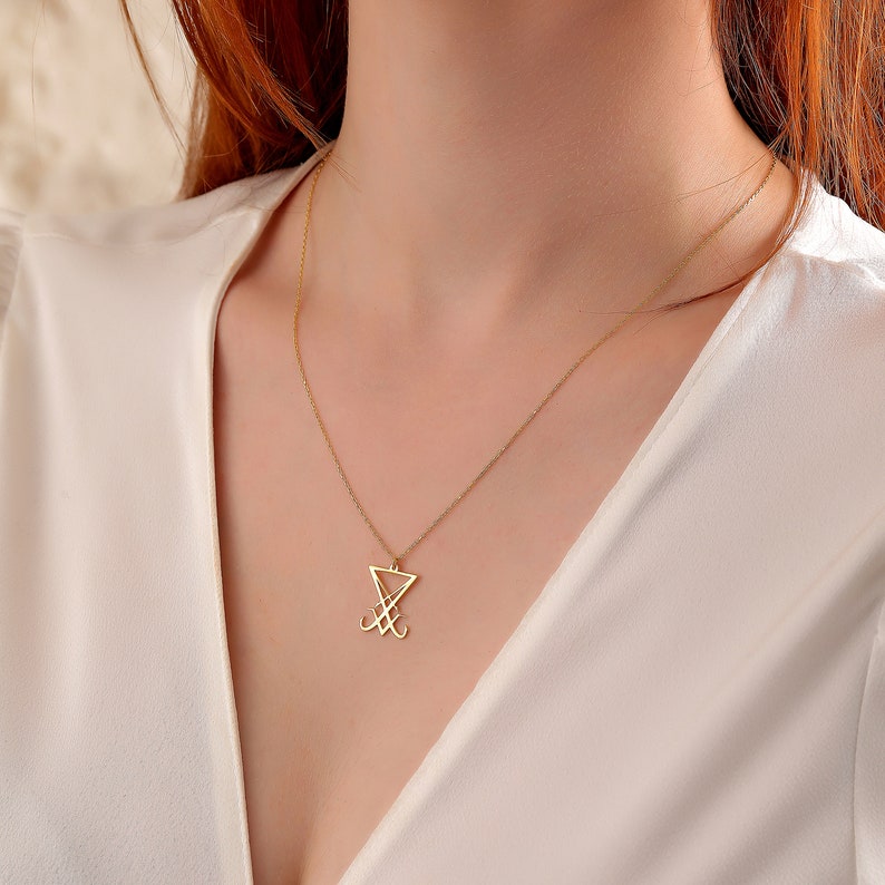 K&ouml;nnte beinhalten: Eine goldene Kettenkette mit einem Anh&auml;nger in Form eines stilisierten Dreiecks mit einem Symbol darin. Der Anh&auml;nger ist ein beliebtes Symbol in okkulten und esoterischen Traditionen.