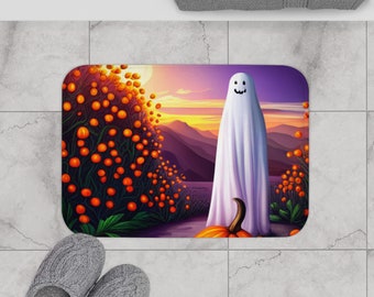 Puede incluir: Una alfombra de baño con temática de Halloween con un fantasma blanco de pie frente a una calabaza y un campo de calabazas naranjas. El fondo es una puesta de sol púrpura con montañas en la distancia.