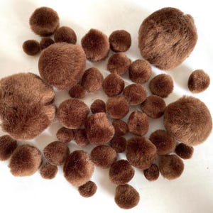 May include: A collection of brown, fluffy pom-poms of varying sizes scattered on a white surface. The pom-poms have a soft, textured appearance, suggesting they are made of a plush material. Ideal for craft projects.