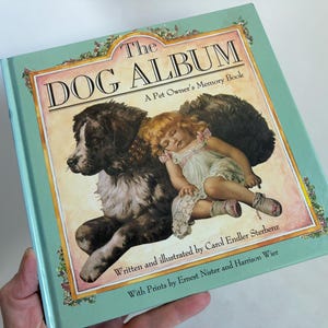 May include: A hardcover book titled The Dog Album: A Pet Owner's Memory Book with a light green cover. The front features an illustration of a black and white dog lying next to a sleeping child in a white dress, with text by Carol Endler Sterbenz.