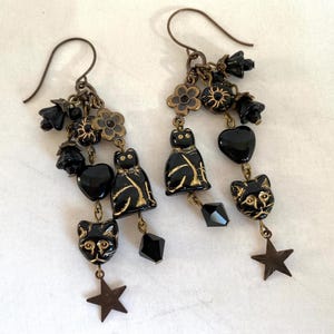 May include: A pair of dangle earrings featuring black cat charms with gold accents, black heart and star charms, and floral and bell-shaped accents. The earrings have a vintage, gothic aesthetic.