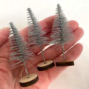 May include: Three small silver glitter Christmas trees with brown wooden bases.