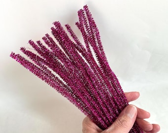 May include: A bundle of magenta pipe cleaners, held in a hand, against a white background. The pipe cleaners are made of a fuzzy, glittery material, and are bunched together. The image is well-lit and the colors are vibrant.
