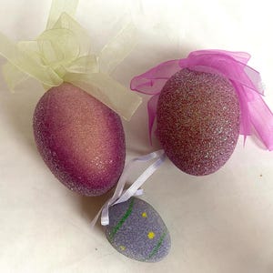 May include: Three decorative Easter eggs. Two are glittery purple with sheer ribbon bows. The third is a smaller, lavender egg with yellow dots and green stripes. All are on a white surface.