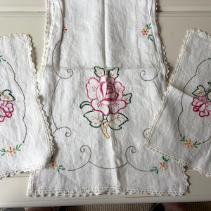 May include: A white linen table runner and two matching rectangular placemats featuring delicate hand-embroidered pink rose motifs with green leaves and small yellow flowers. Each piece is finished with a decorative white crocheted lace edge.