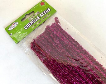 May include: A package of pink glitter chenille stems, also known as pipe cleaners. The package is labeled "Glitter Chenille Stems" and "Cure-pipes brillants".
