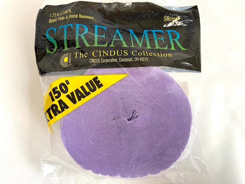 May include: A roll of lavender streamer paper in a clear plastic package. The package has a black top with the word "STREAMER" in green and blue letters. A yellow tag reads "150' EXTRA VALUE". The streamer is 1.75 inches wide and 150 feet long.