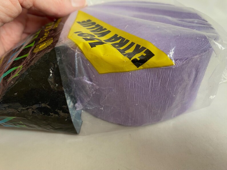 May include: A roll of lavender crepe paper is partially visible inside a clear plastic package. A yellow label with the words "EXTRA VALUE" in black is attached to the package. The paper has a textured surface.